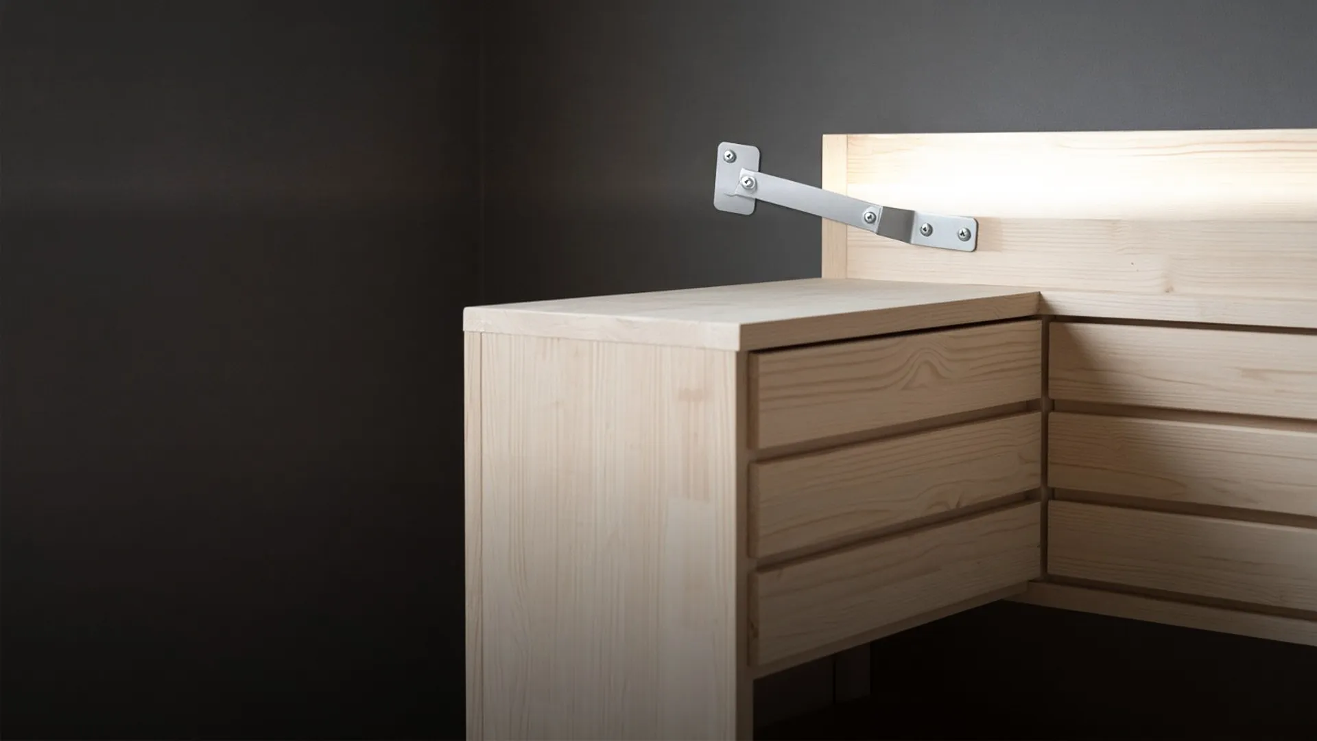 A plain metal bracket and strap joining the back of a dresser to a wall