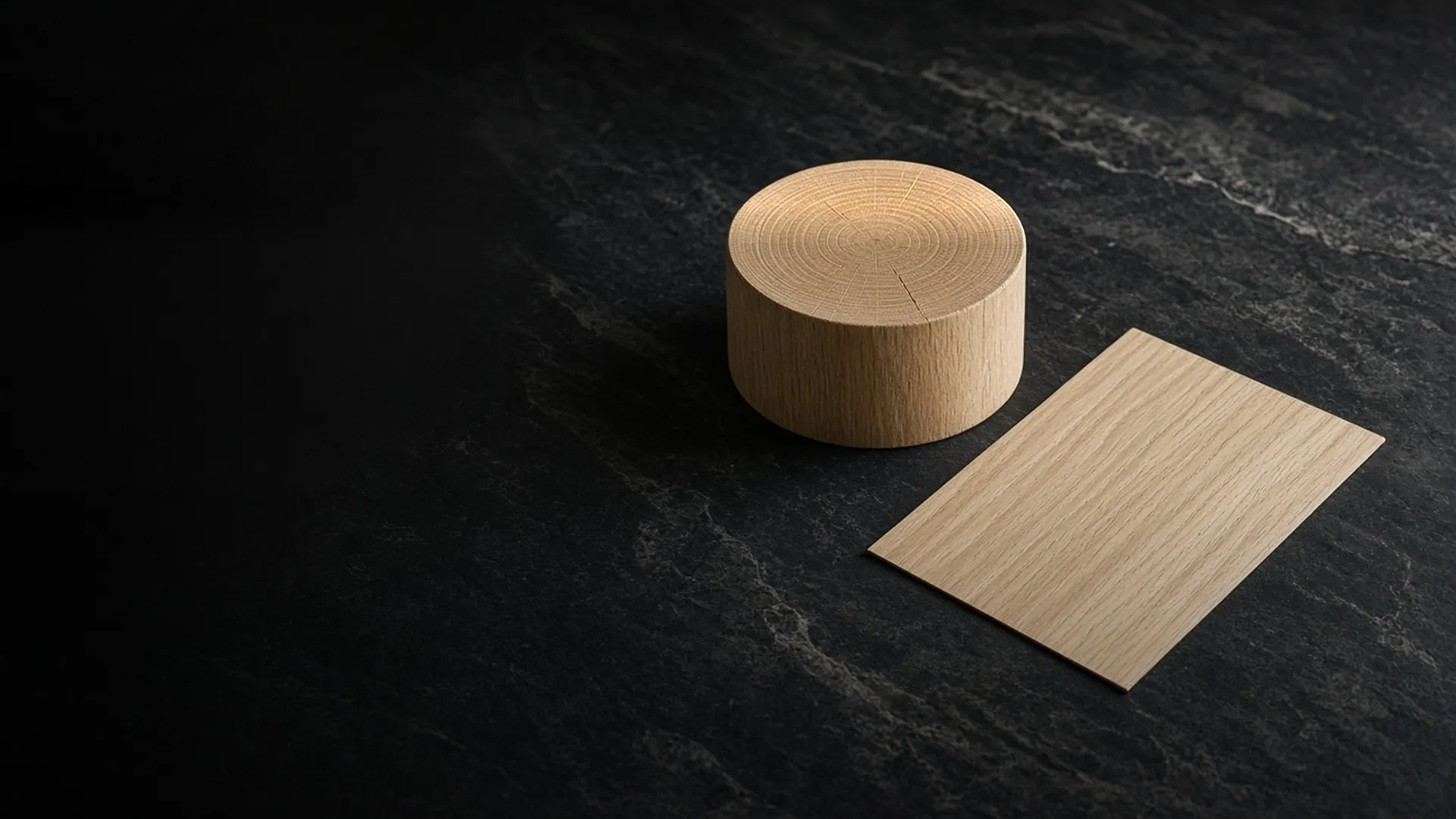 A round bare wood block beside a thin flat sheet of wood veneer on dark stone